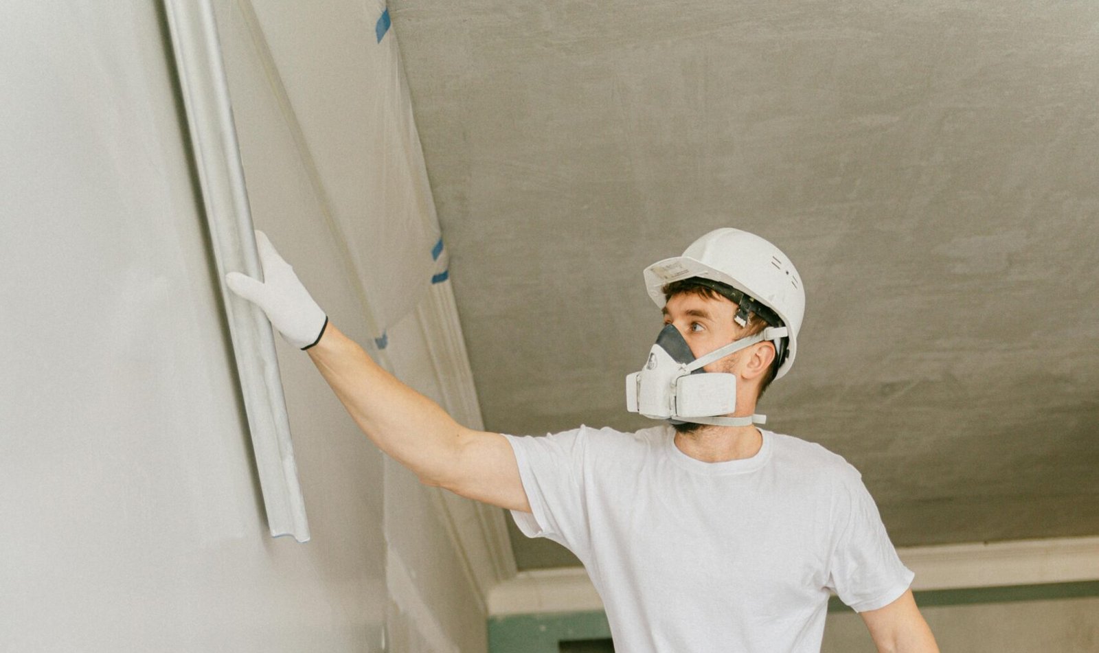 Construction worker on stilts painting a wall indoors, wearing protective gear and a hard hat.